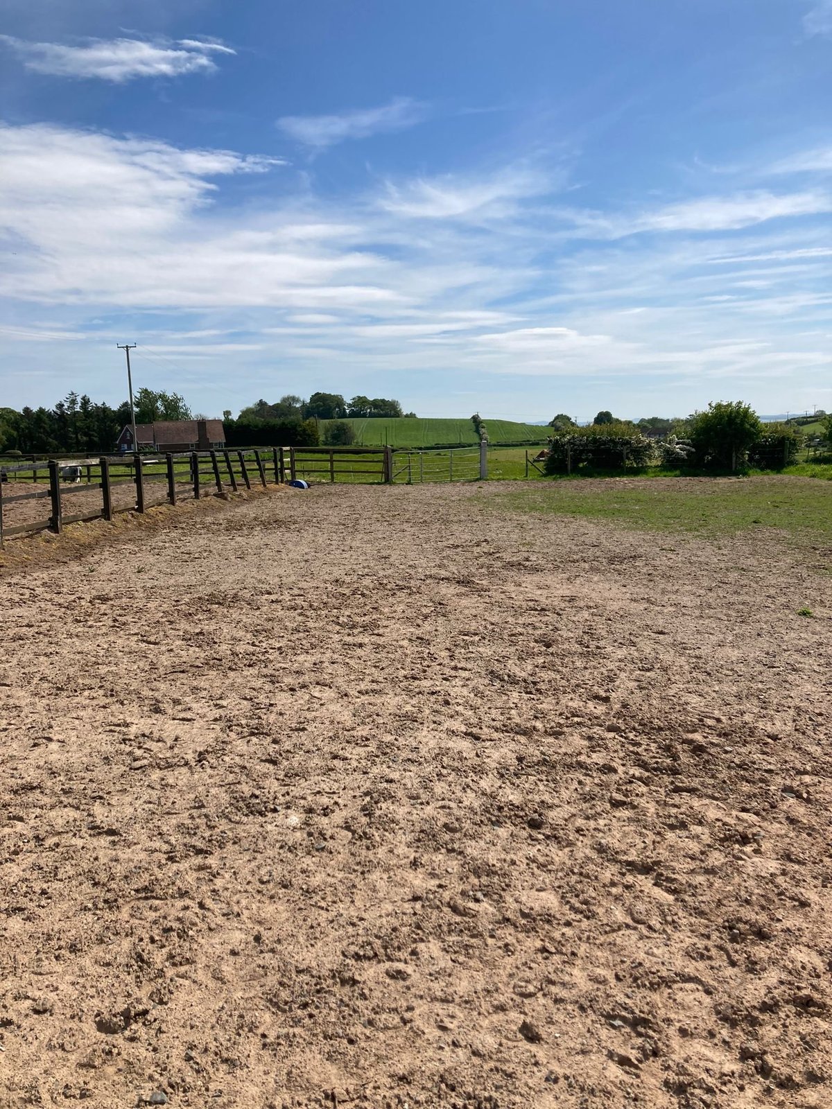 A large dirt ranch or livestock enclosure with wooden fencing, green pasture fields, trees, and a clear blue sky with white clouds