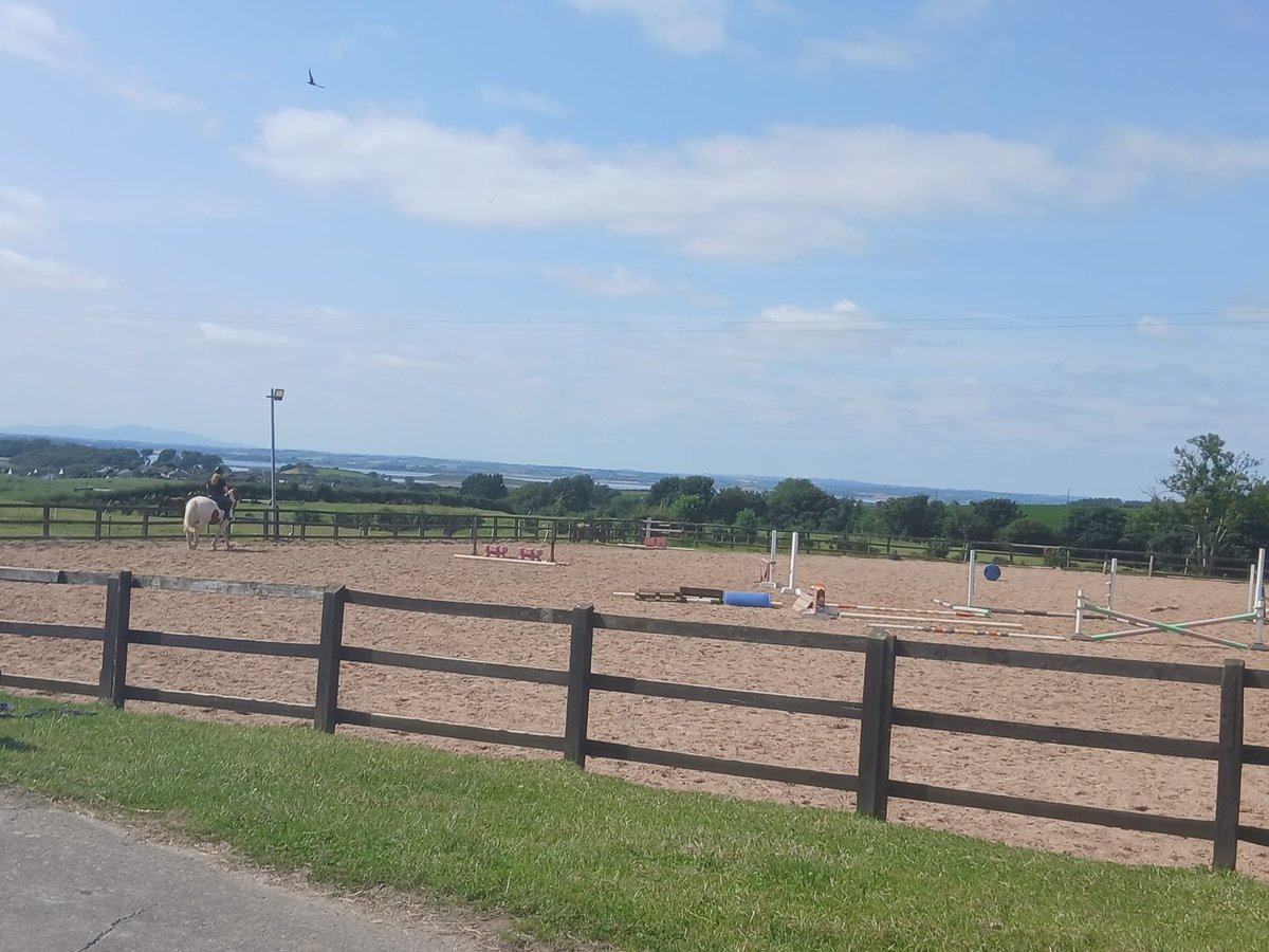 Wooden fence bordering a large empty dirt riding arena with green pastures and countryside landscape in the background