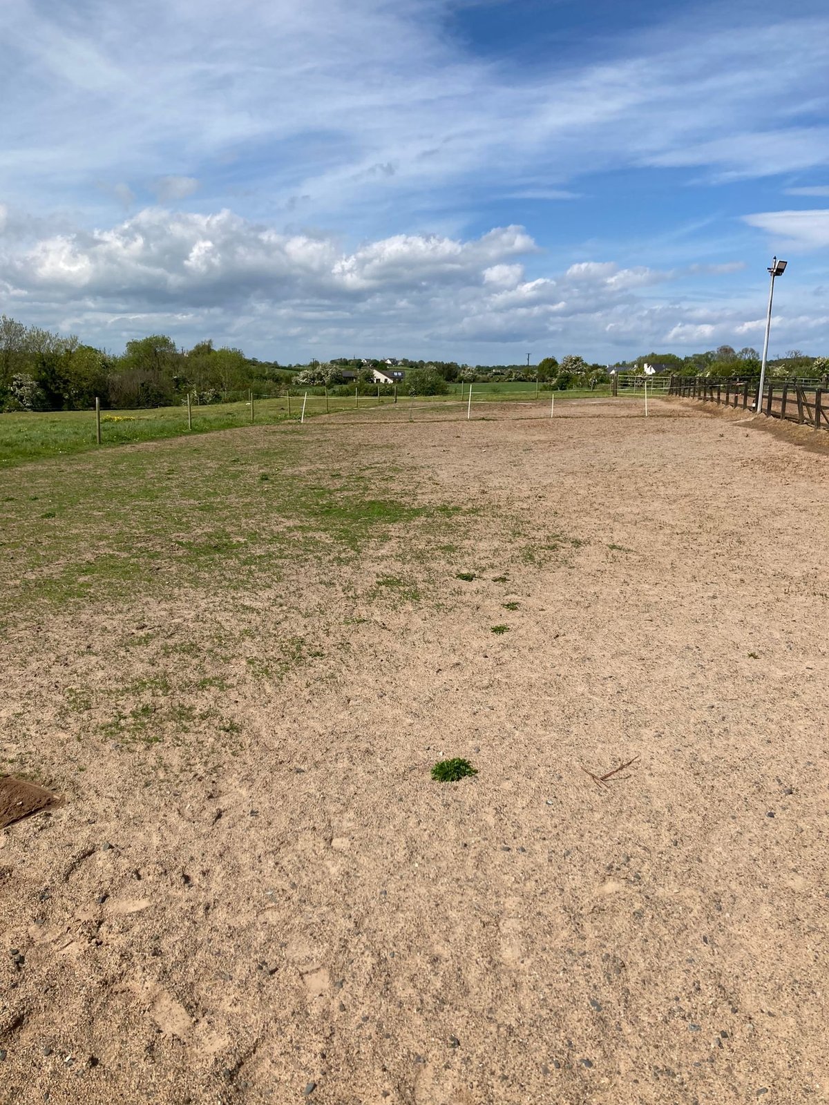 Empty dirt horse arena with fencing, green pasture, and houses visible under blue sky with white clouds