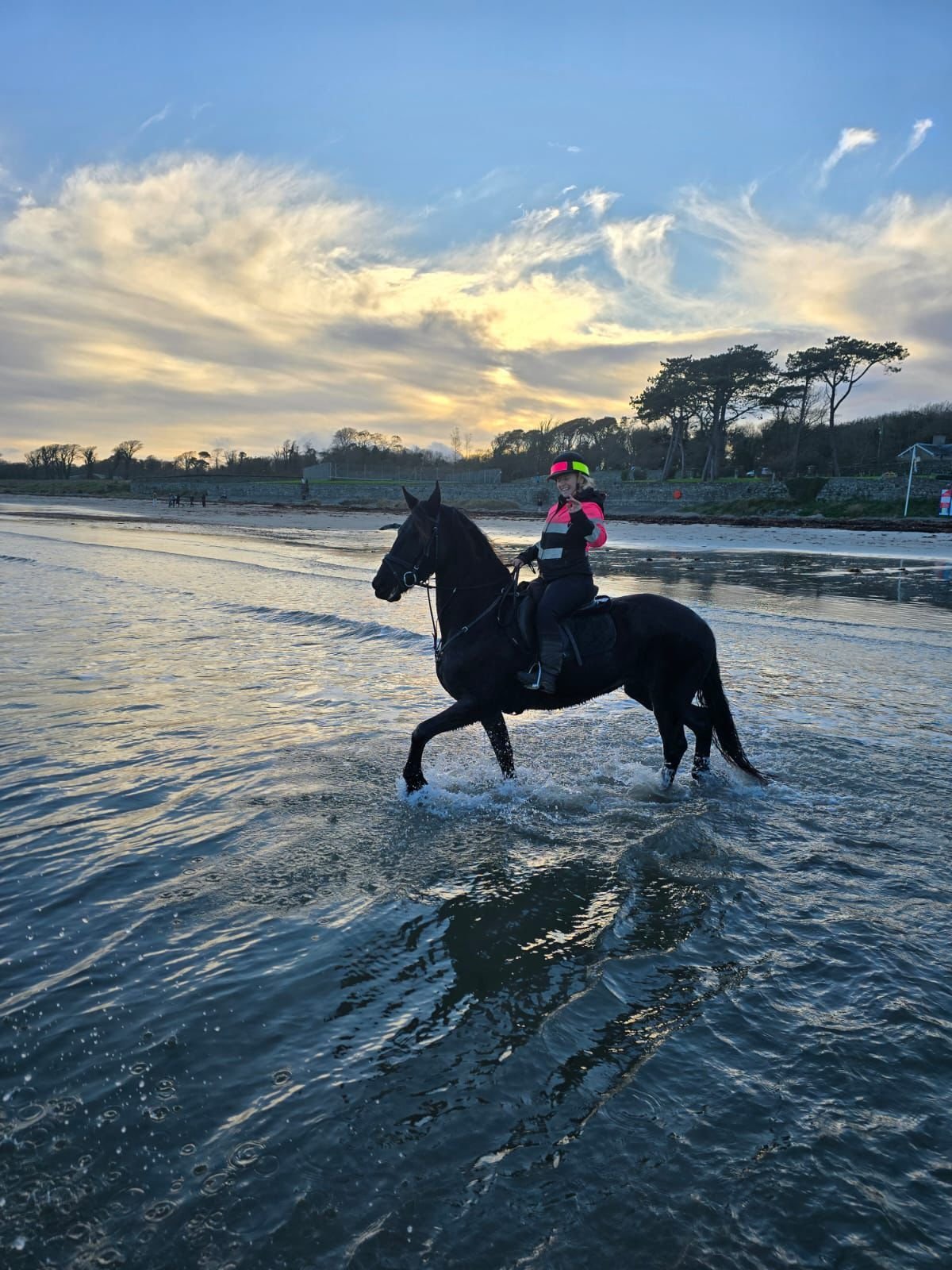 A person in pink riding a black horse through shallow coastal water at sunset with blue sky and clouds above.