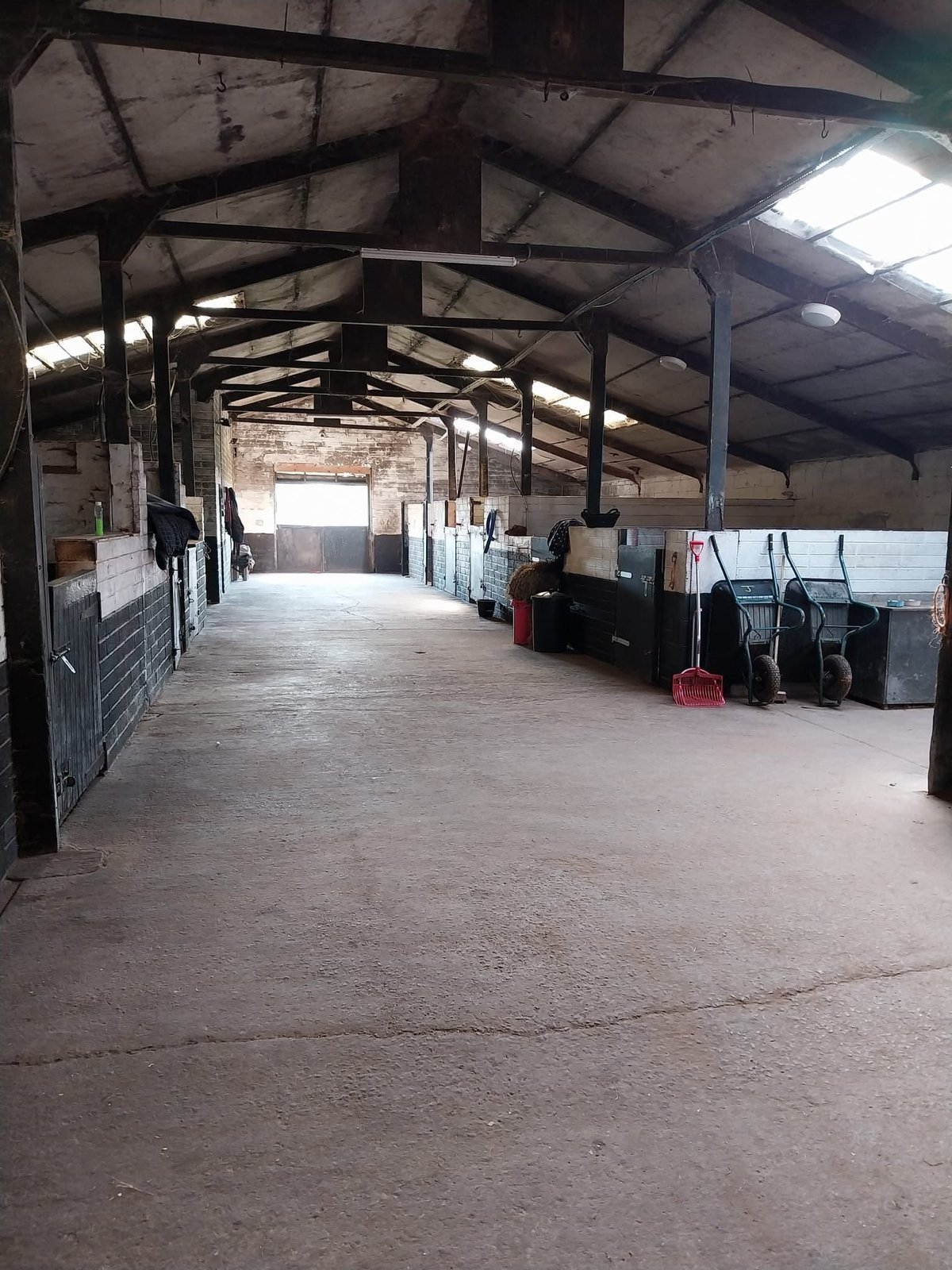 Interior of a large industrial warehouse with metal beam ceiling, dirt floor, and scattered equipment and containers.