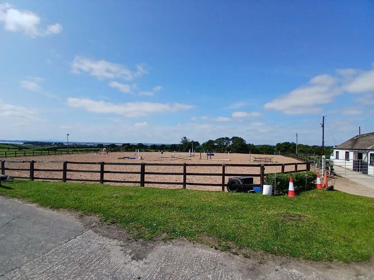 Rural farm scene with wooden fence enclosure, green fields, blue sky with clouds, and farm buildings in background